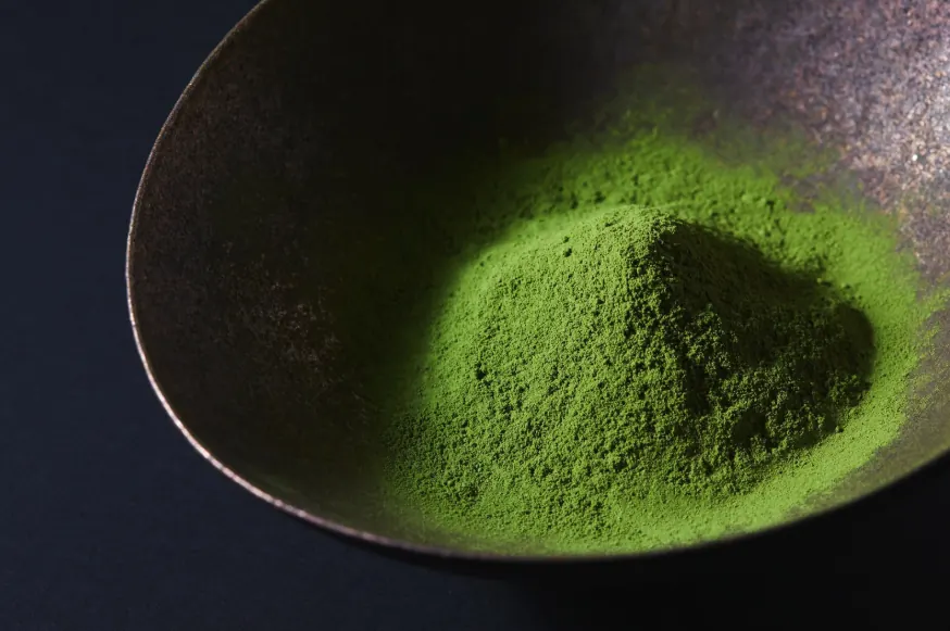 Mound of fine, vivid green matcha powder in a metal dish against a dark background.
