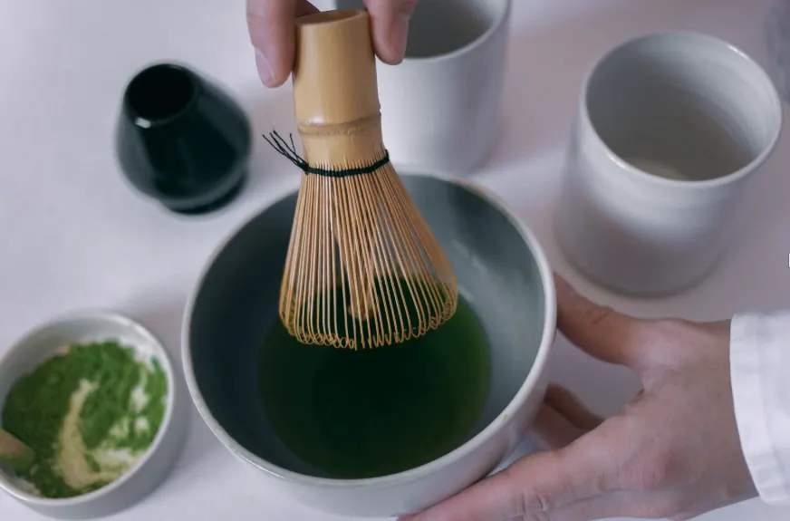 Hands whisking matcha with a bamboo whisk in a ceramic bowl, with a dish of sifted matcha powder alongside.