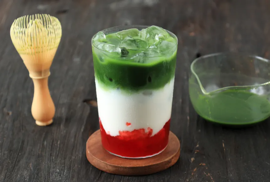 Layered iced strawberry matcha latte on a wooden coaster beside a bamboo whisk and a bowl of whisked matcha.