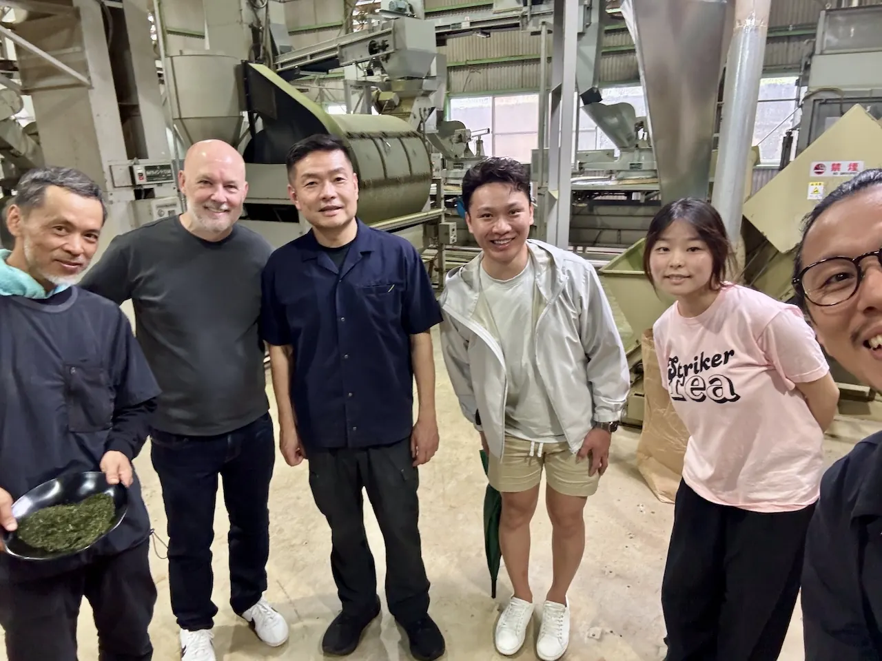 Cafe buyers standing with a producer inside a Japanese tea processing factory, one holding a tray of fresh leaf.