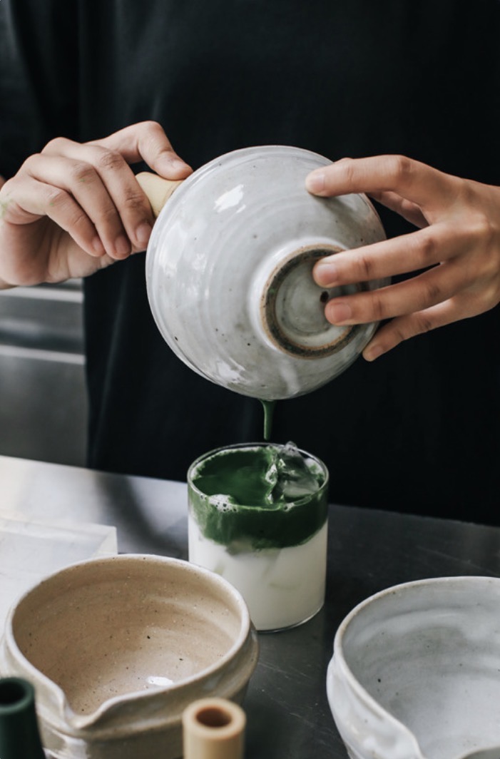 A barista pours whisked matcha from a bowl into a glass of iced milk.