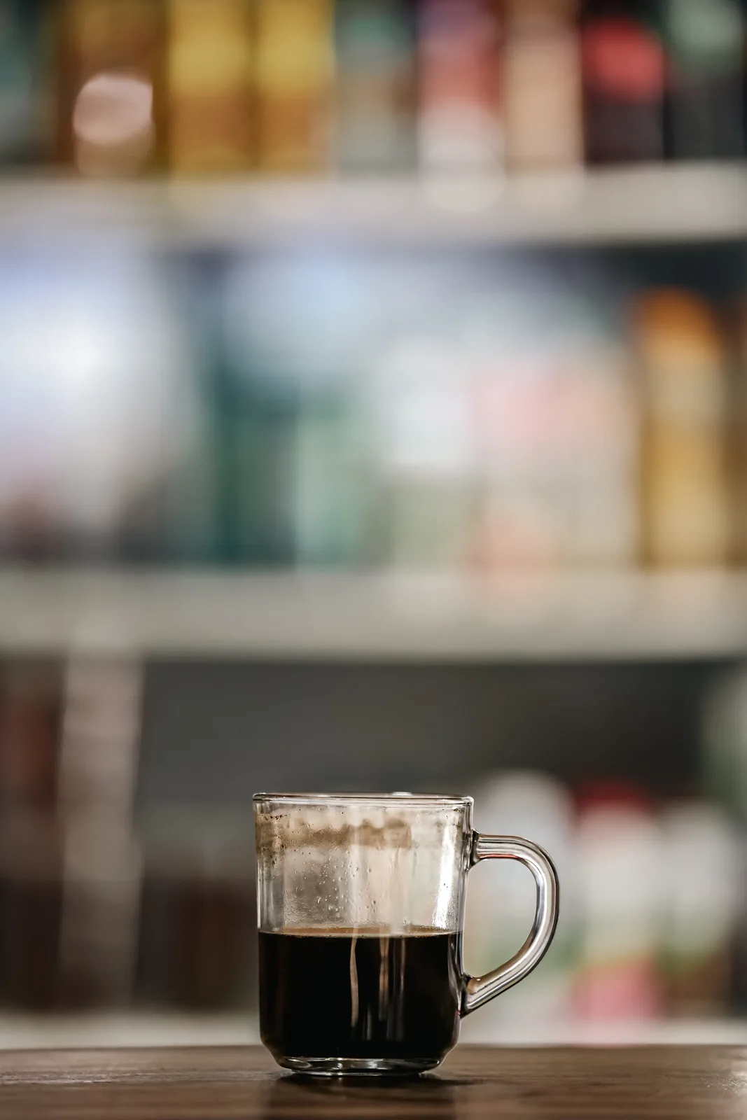 A glass coffee cup on a wooden table with a display refrigerator blurred in the background.