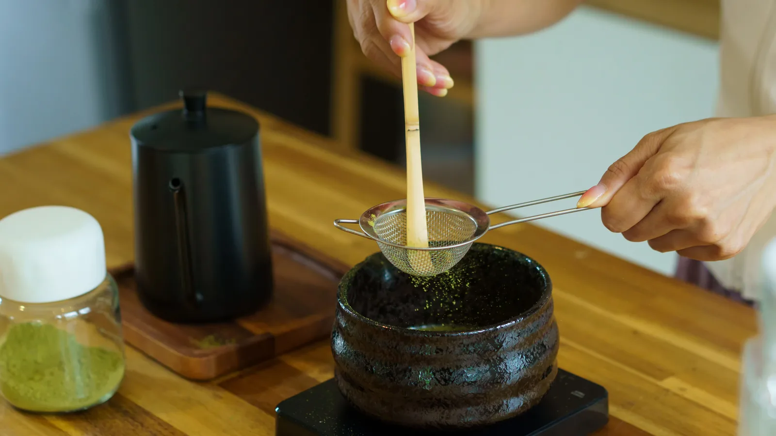Sifting matcha powder into a ceramic bowl with a bamboo scoop and a fine mesh strainer.