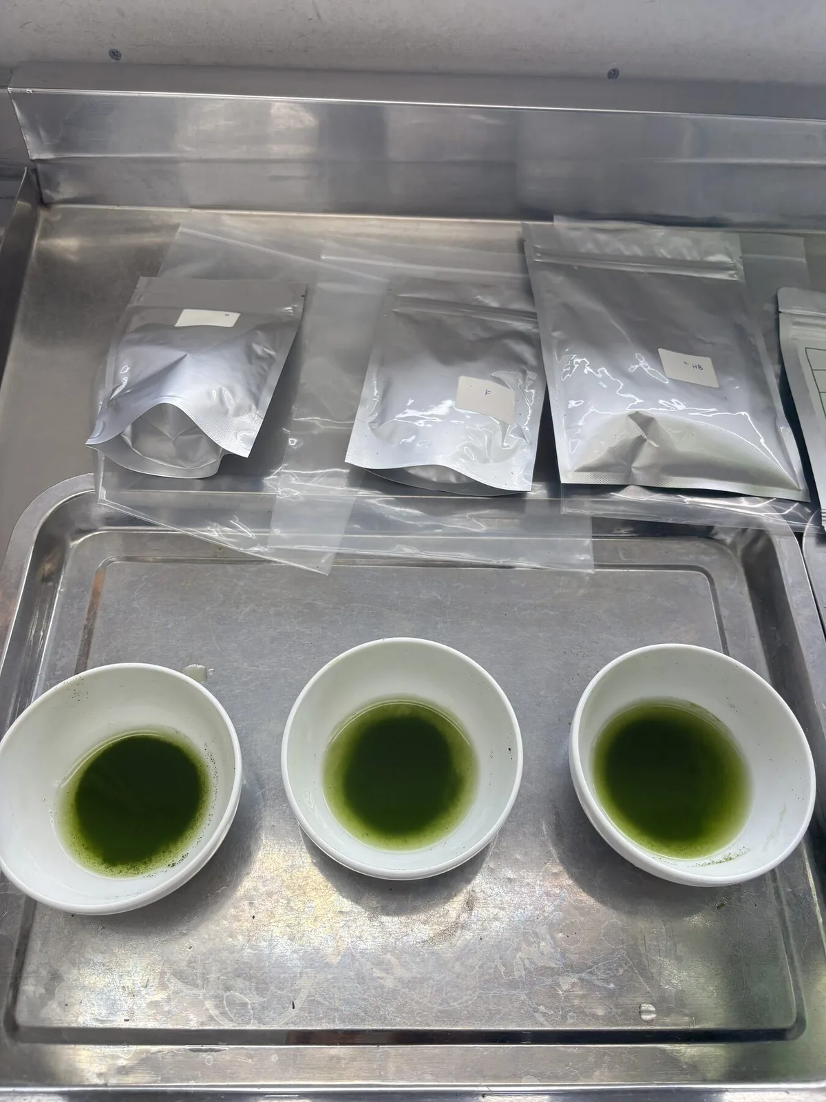 Three white bowls of whisked matcha and sample bags on a tray for cupping.