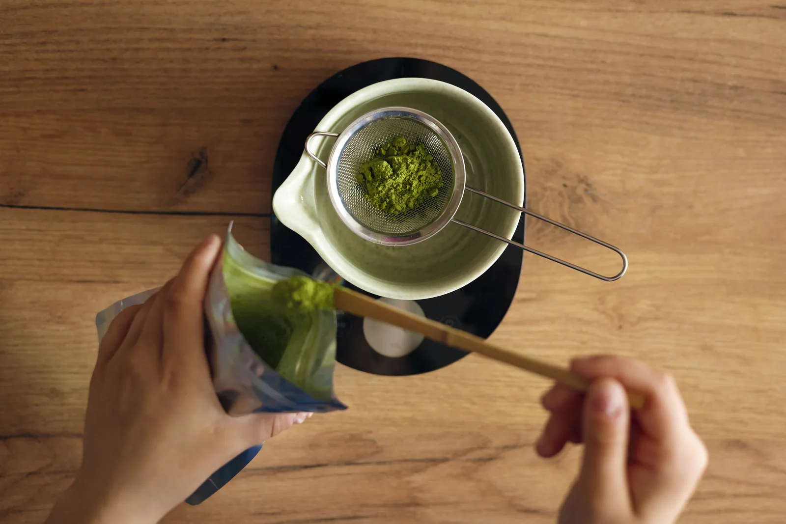 Hands sifting matcha powder through a fine sieve into a small pitcher before any liquid is added.