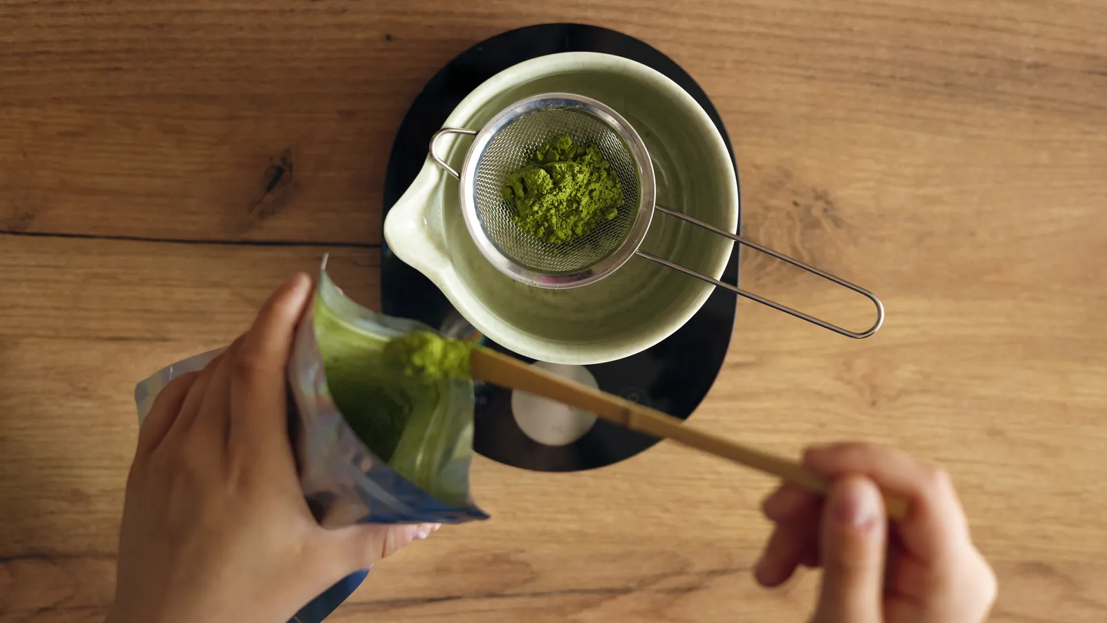 Hands sifting matcha powder from a sample pouch through a fine strainer, seen from above.