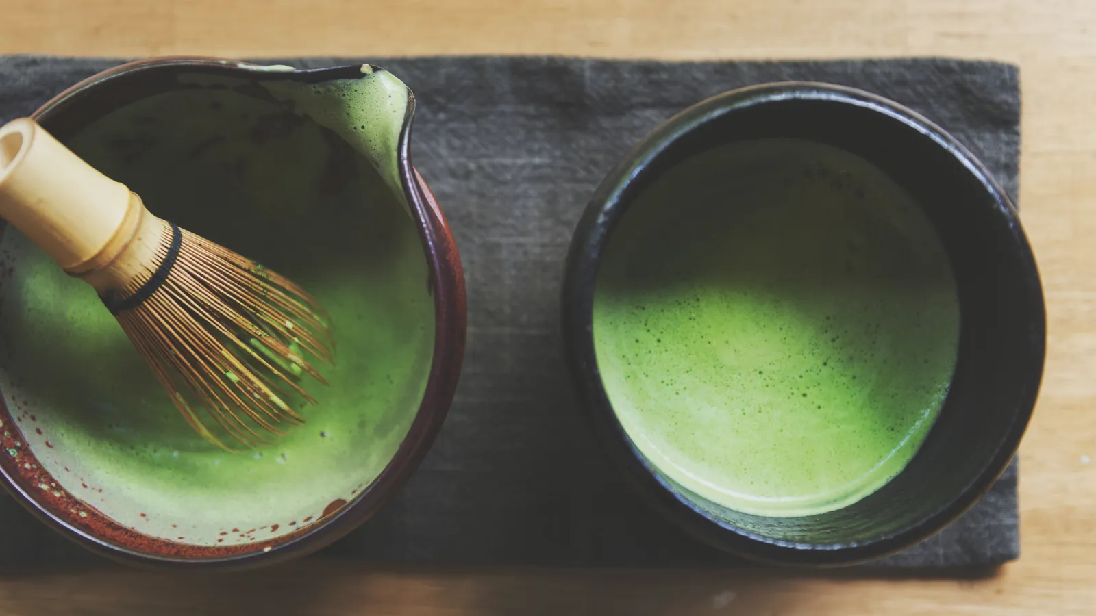 A bamboo whisk resting in one bowl of matcha beside a second bowl of whisked tea.