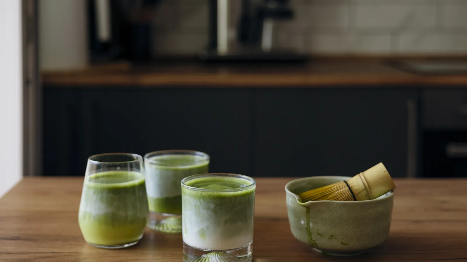 Three glasses of matcha of different strengths on a counter beside a bowl and whisk.