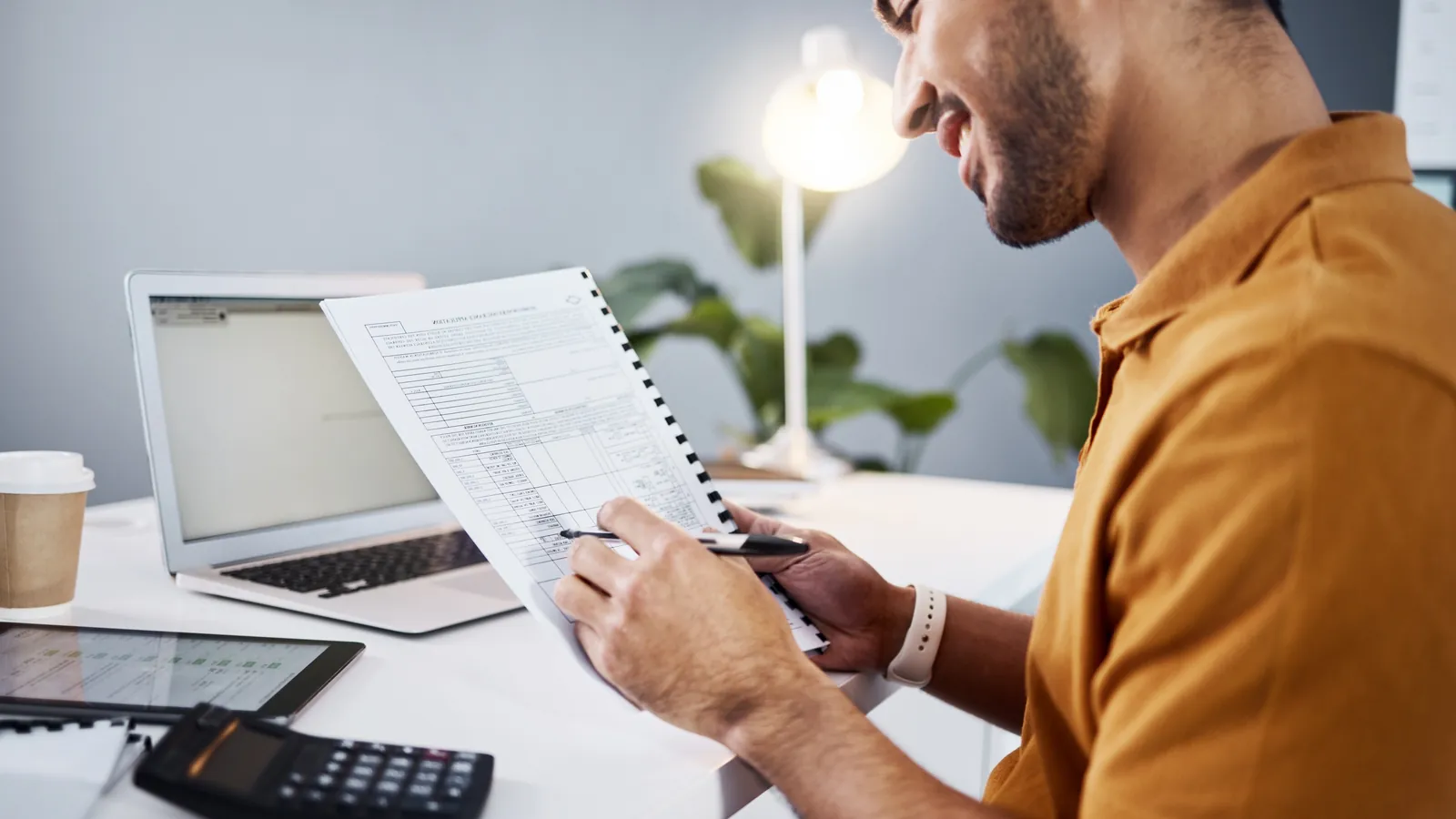 A cafe owner going through an invoice at a desk with a calculator and a laptop.