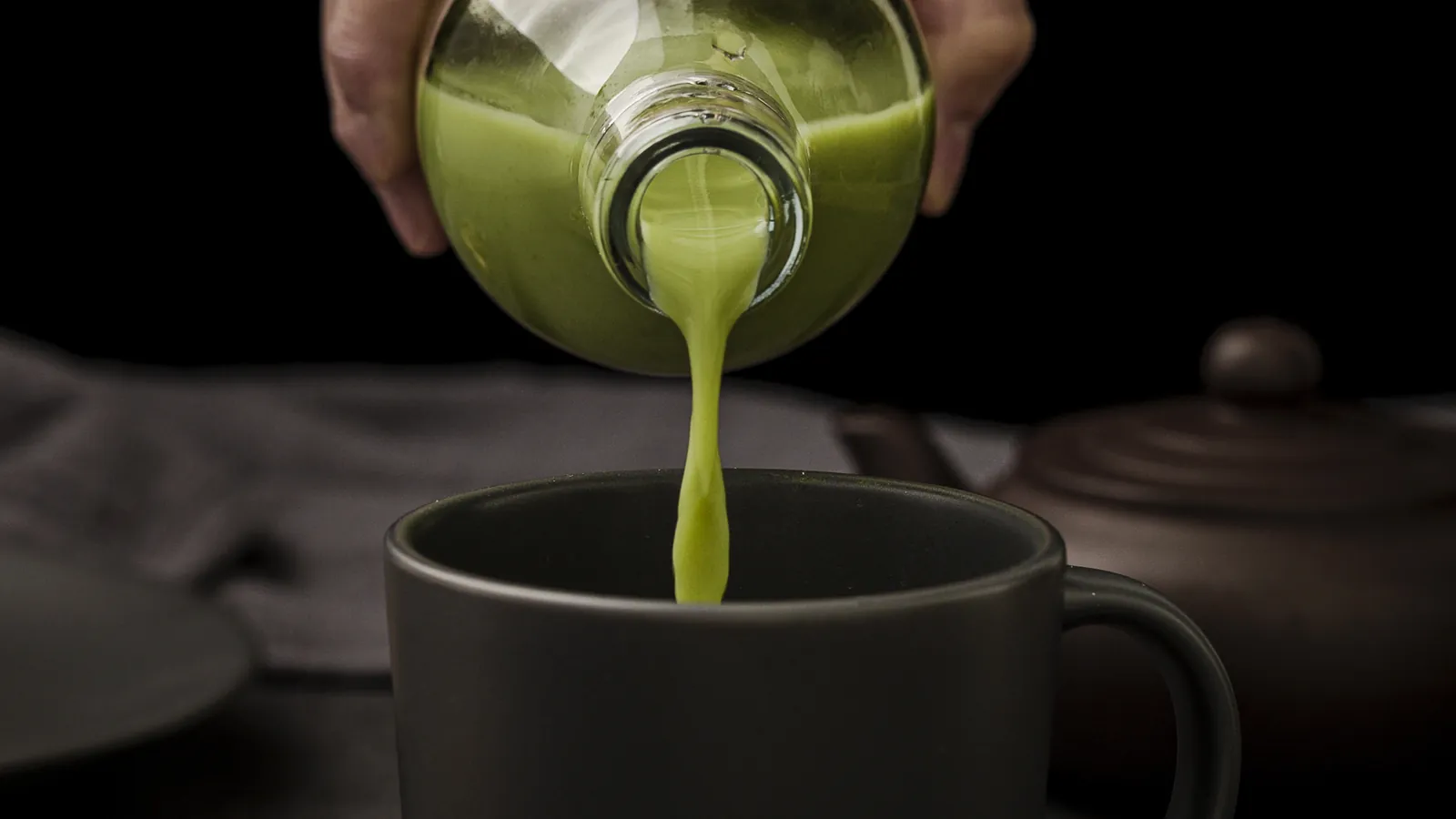 Thick whisked matcha being poured into a dark mug against a black background.