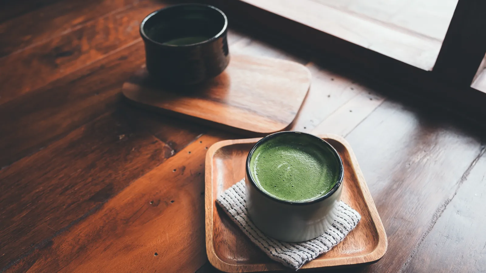 Two servings of hot matcha on wooden trays in warm low light.
