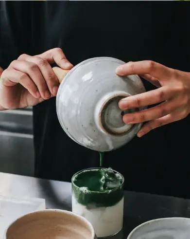 Barista pouring whisked matcha from a spouted bowl over iced milk to build a layered matcha latte.