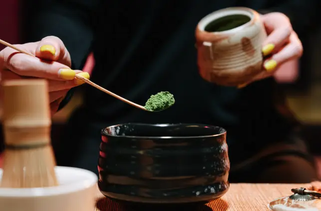 Matcha being measured with a bamboo scoop into a dark tea bowl, with a whisk and caddy alongside.
