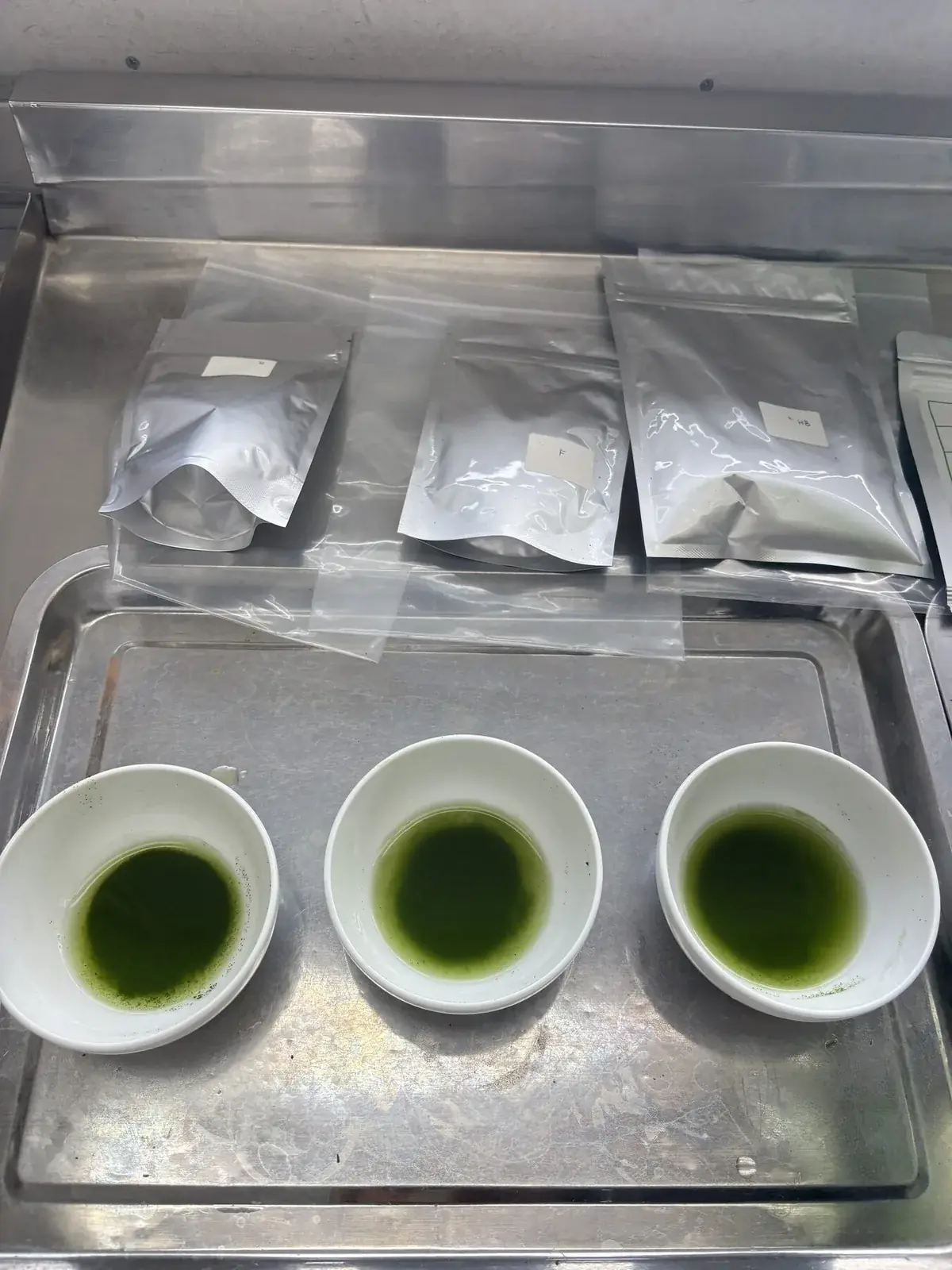 Three white bowls of whisked matcha and sample bags on a tray for cupping.