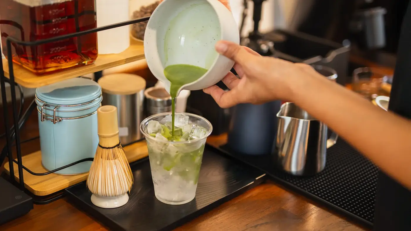 A barista pouring matcha from a bowl into a glass of ice on a cafe bar with tea canisters and a bamboo whisk.