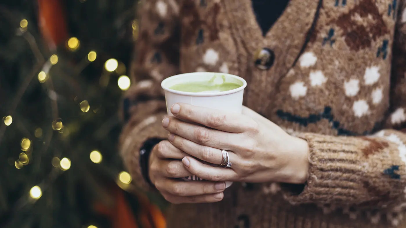 Two hands in a patterned wool cardigan holding a paper cup of hot matcha latte outdoors in winter.
