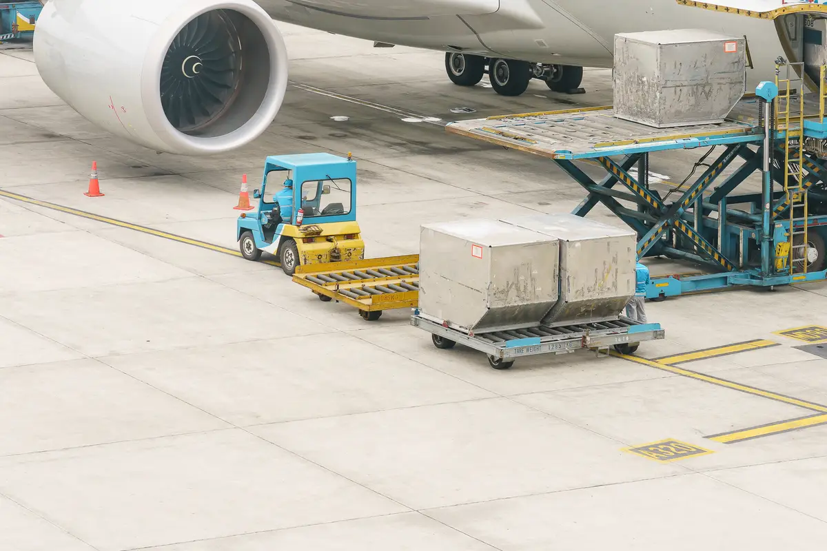 Two aluminium air cargo containers on a dolly beside a wide-body aircraft on the apron, with a loader lifting a third into the hold.