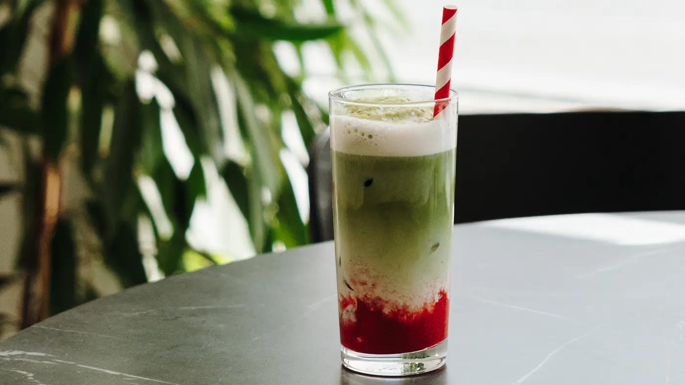 A tall iced matcha latte layered over strawberry, with a red and white paper straw, on a dark table by a sunny window.