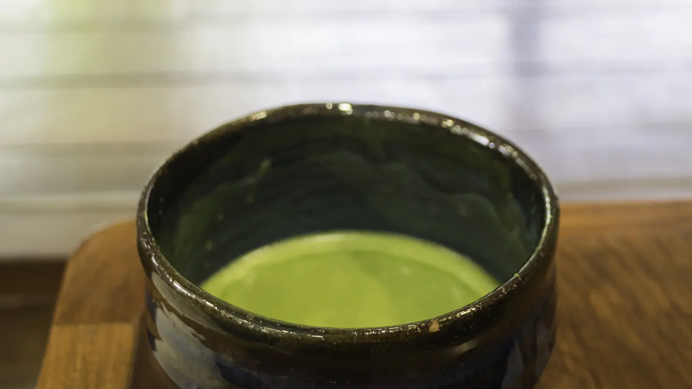 A dark glazed tea bowl of whisked matcha on a wooden tray, against a bright blurred background.