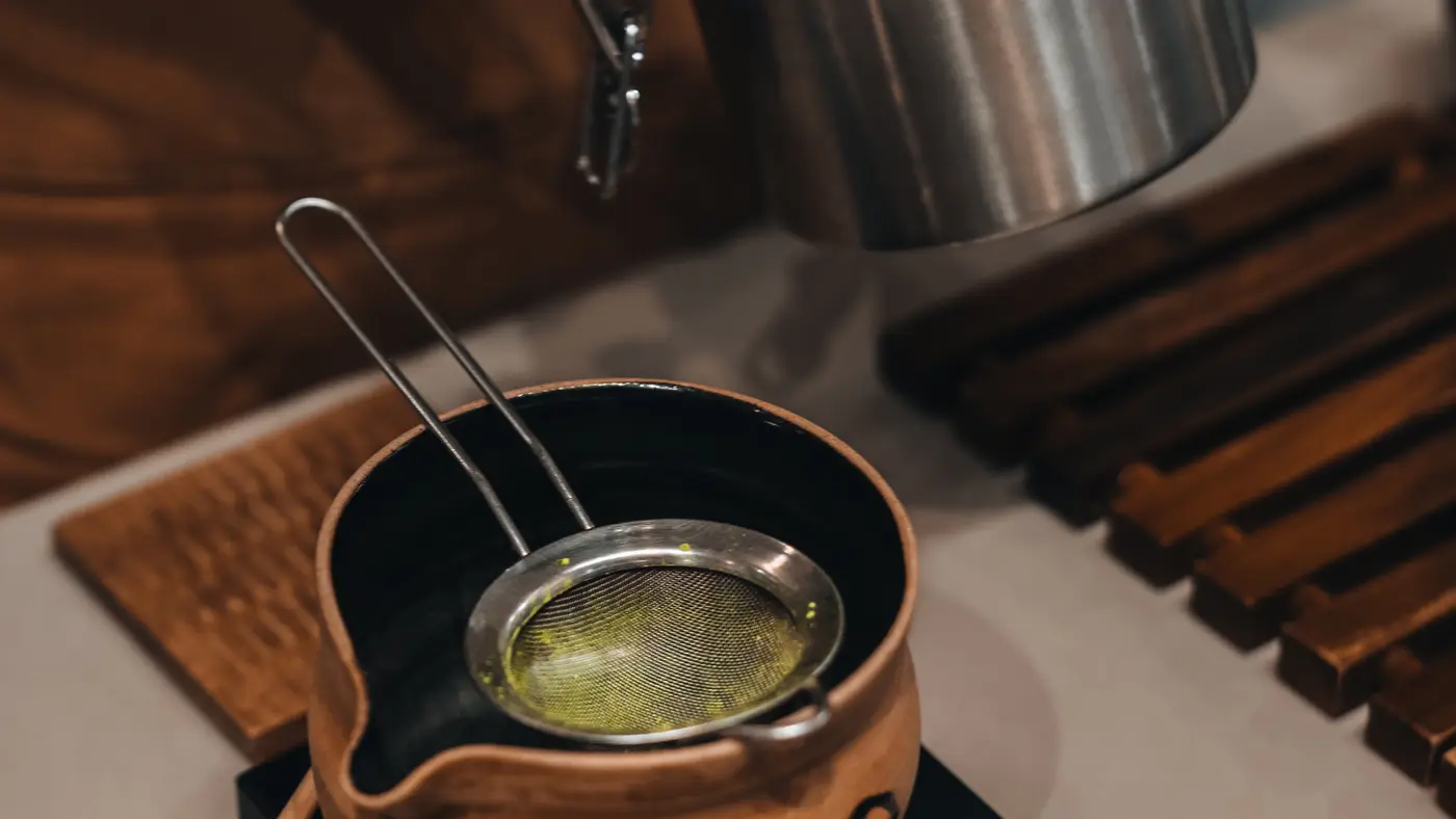 A gloved barista sifting bright green matcha through a steel strainer into a clay pouring bowl on a cafe counter.