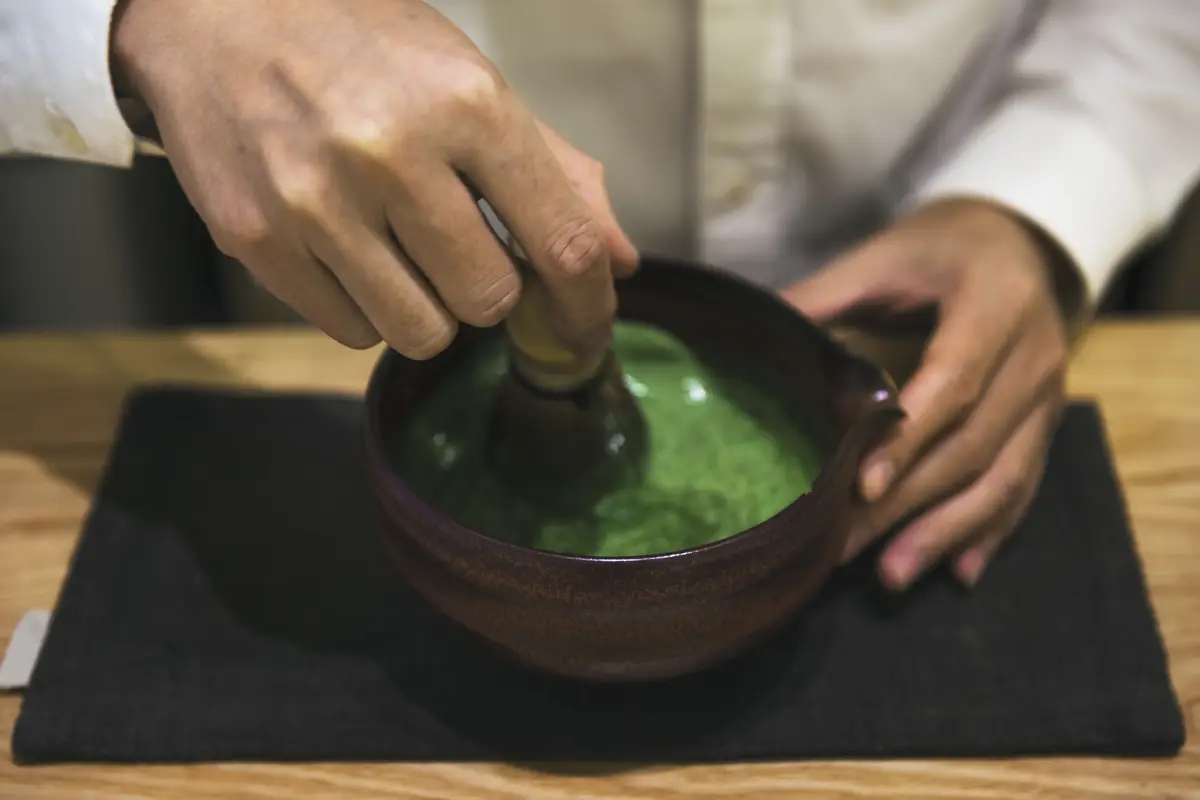 Hands whisking bright green matcha in a dark ceramic bowl on a black cloth mat on a wooden table.