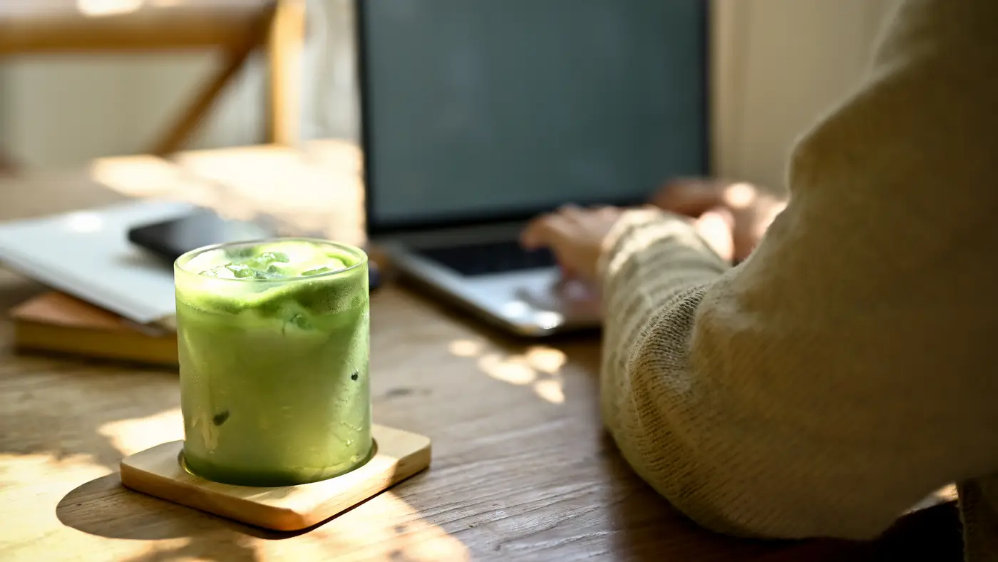 A glass of iced matcha latte on a wooden coaster in strong afternoon sunlight, next to someone working on a laptop.