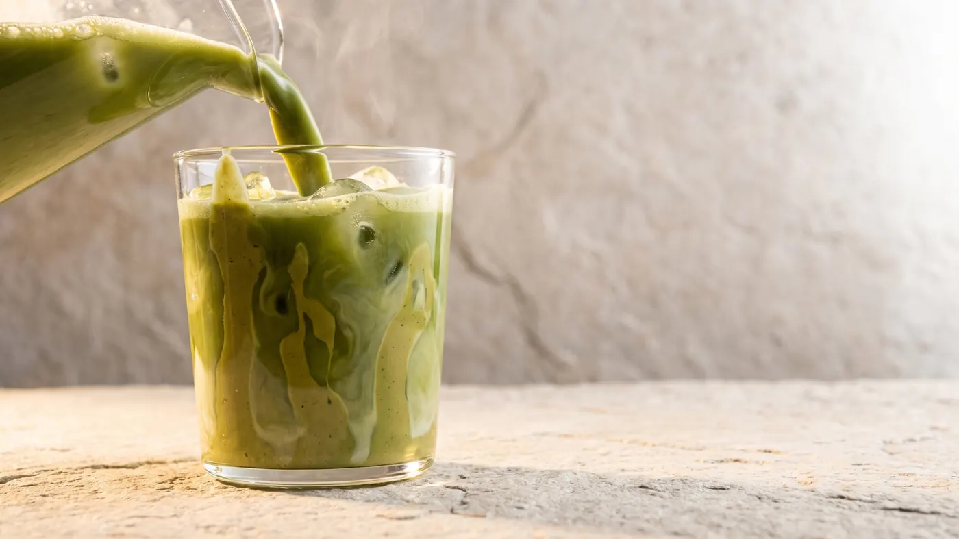 A green iced latte being poured into a glass on a sand coloured stone counter in warm afternoon light.