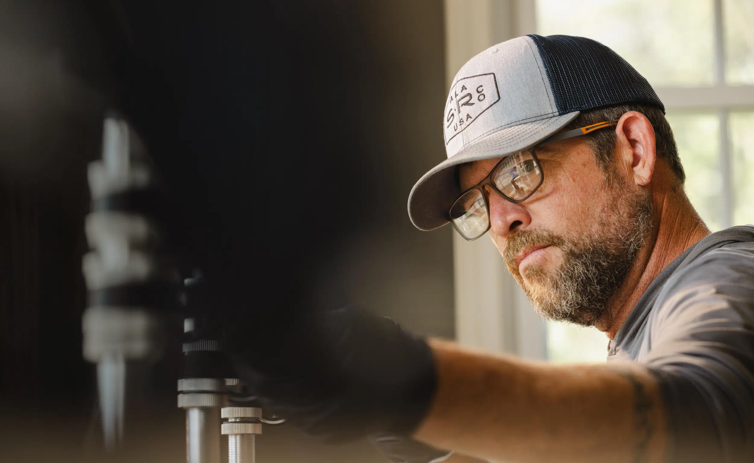 Bearded man wearing glasses and a cap, focused on adjusting equipment with a gloved hand.