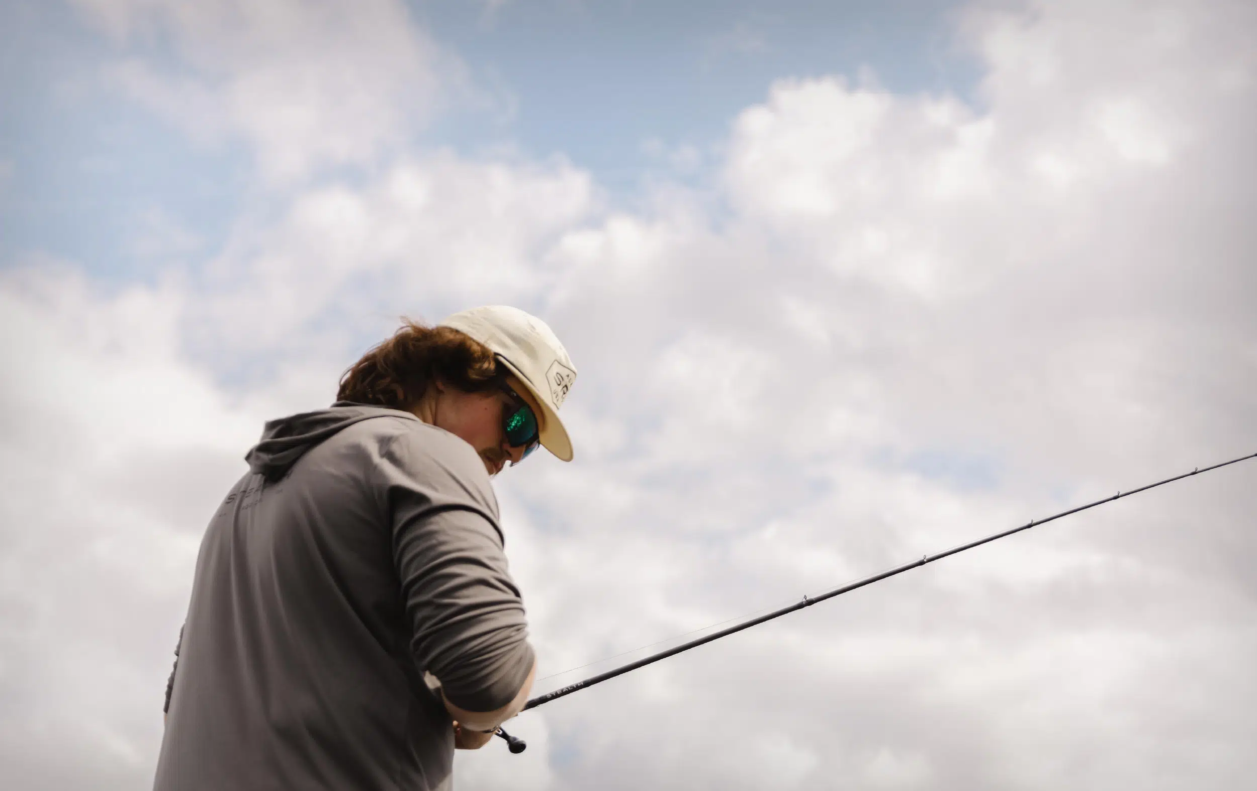 Person wearing a beige cap and sunglasses fishing with a rod under a cloudy sky.