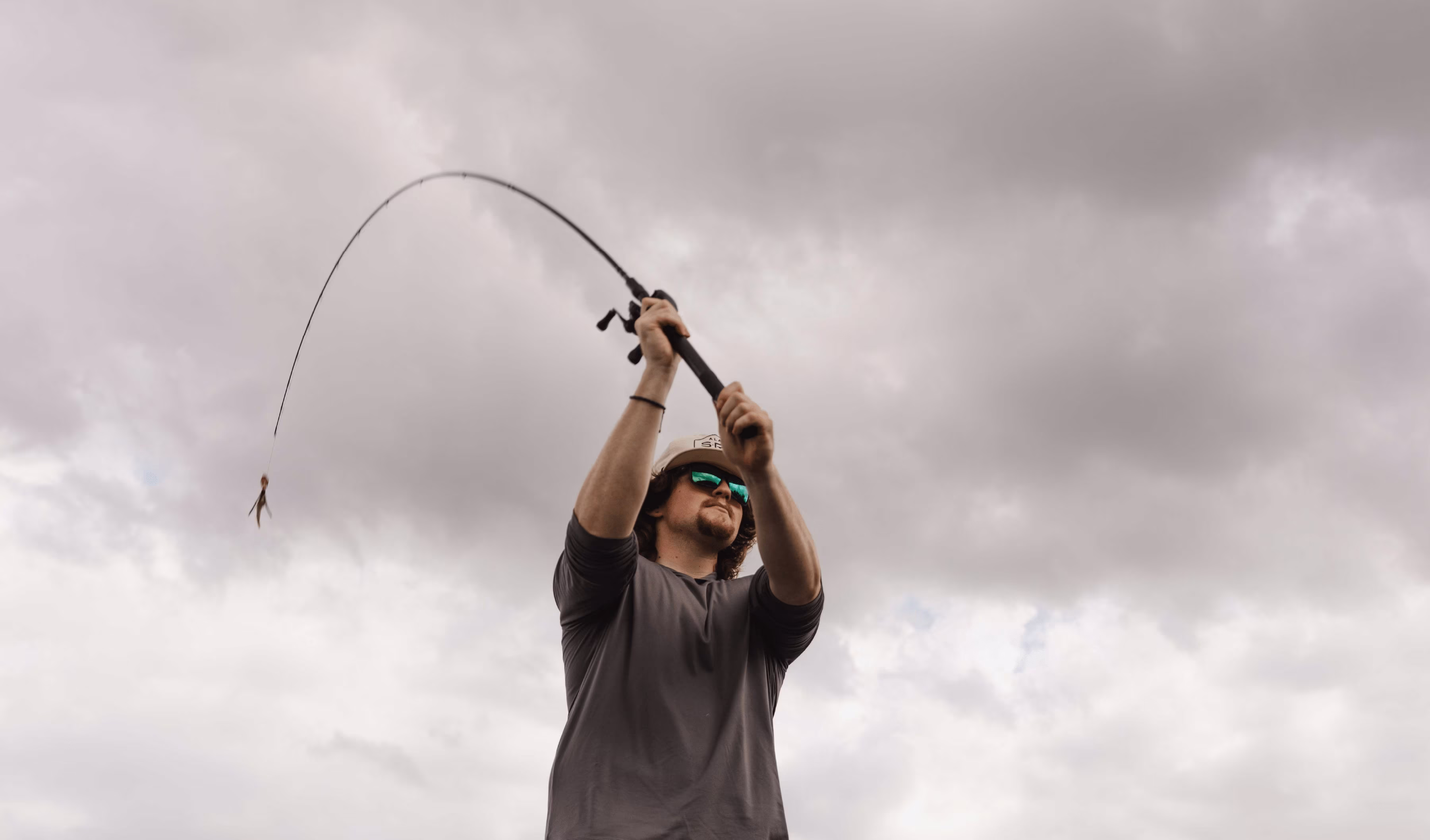 Man wearing sunglasses and a cap casting a fishing rod against a cloudy sky.