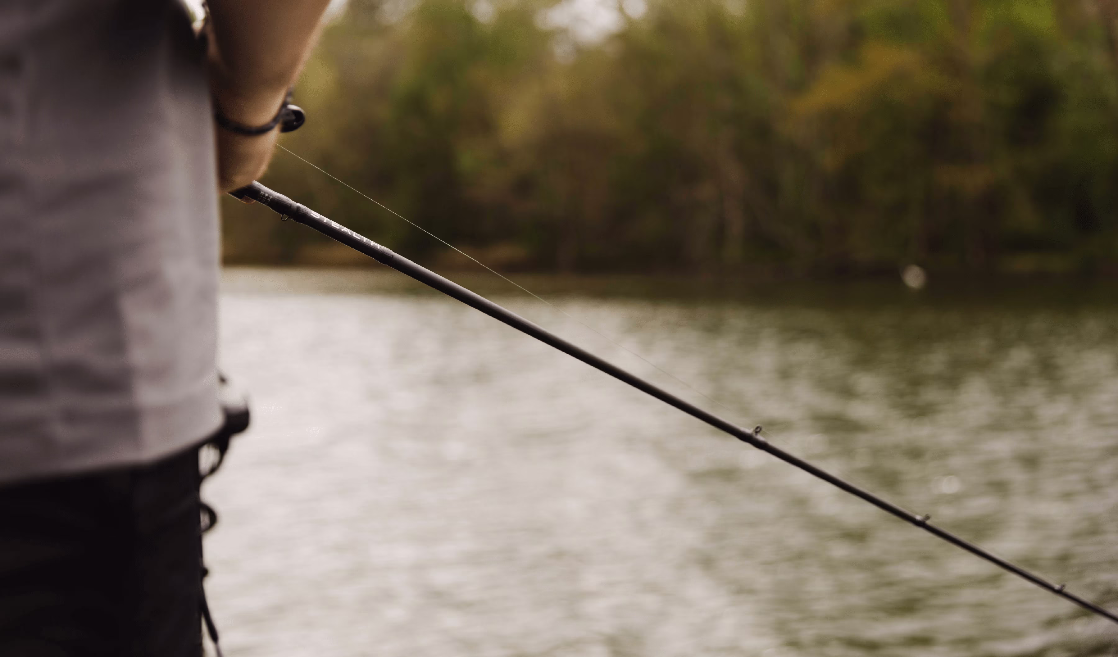 Person holding a fishing rod by a lake with trees in the background.