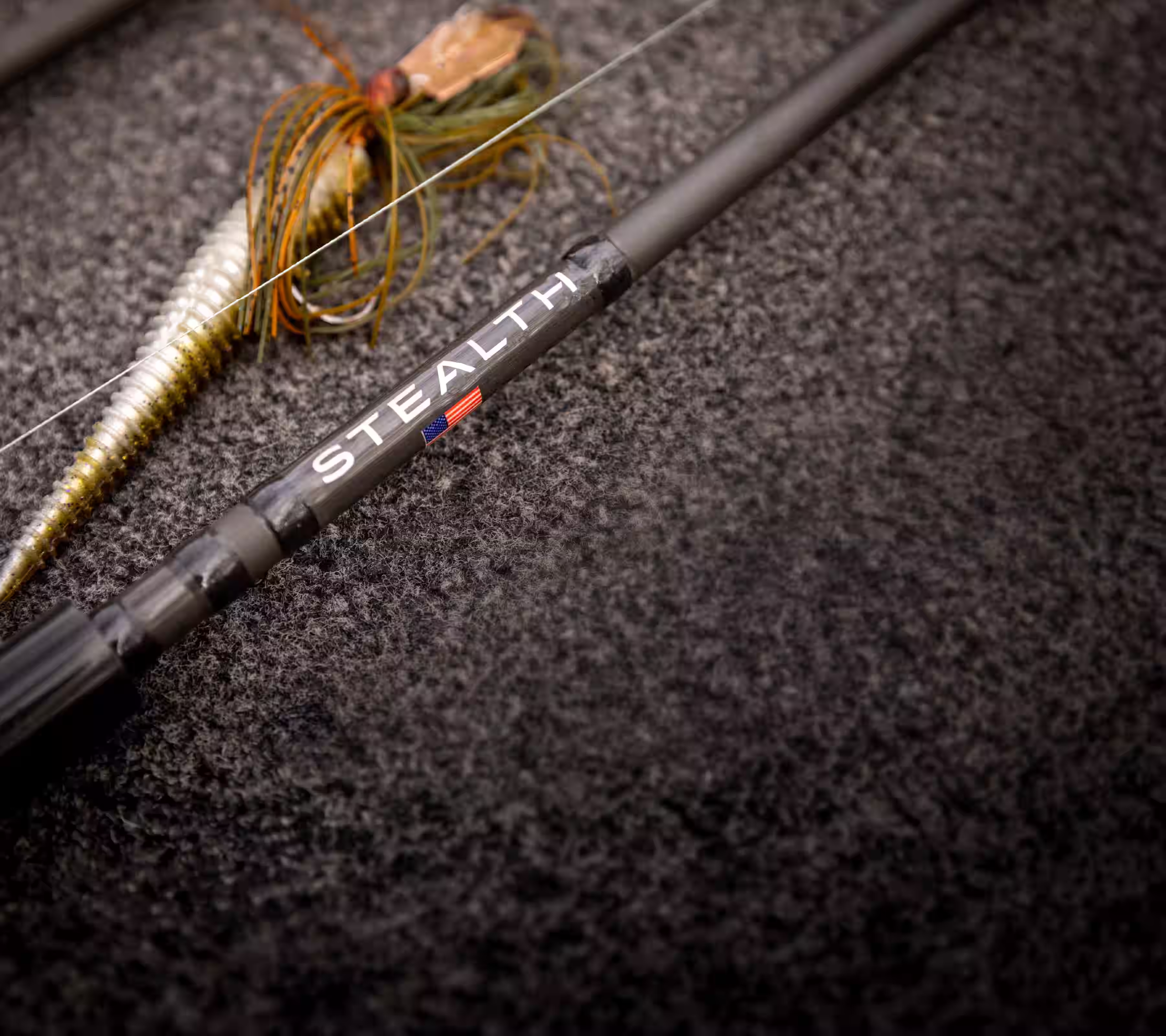 Close-up of a fishing rod labeled 'STEALTH' with an American flag and two fishing lures on a textured surface.