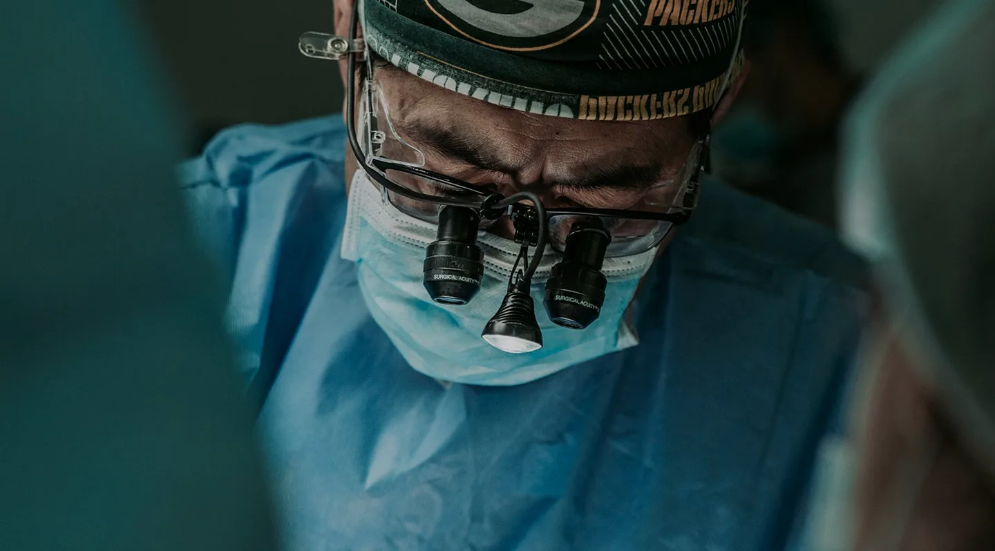 Chirurgien portant un masque, des loupes et une tenue bleue pendant une intervention.