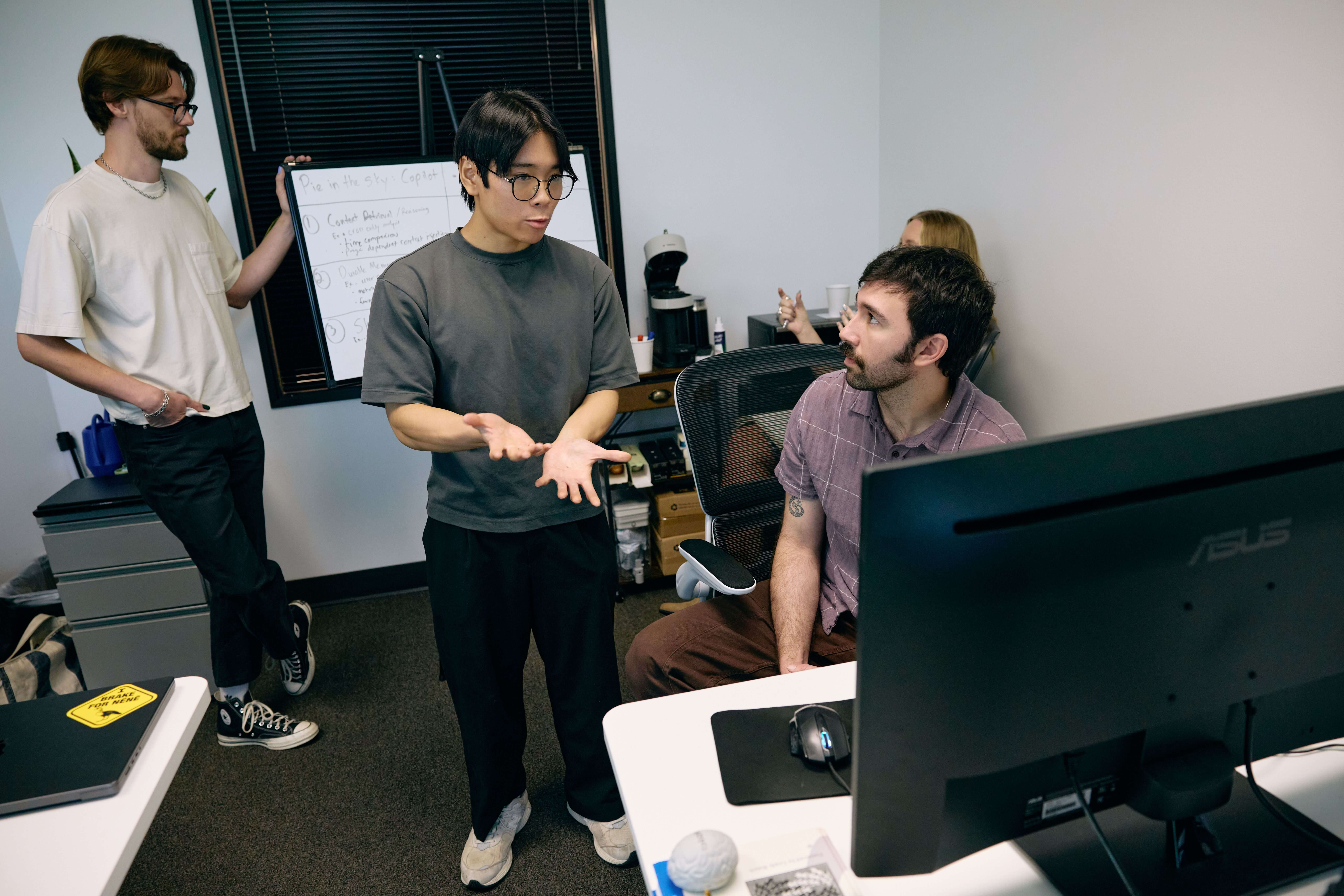 Four colleagues in an office, two discussing by a computer, one writing on a whiteboard, and a fourth visible in the background. Collaborative atmosphere.