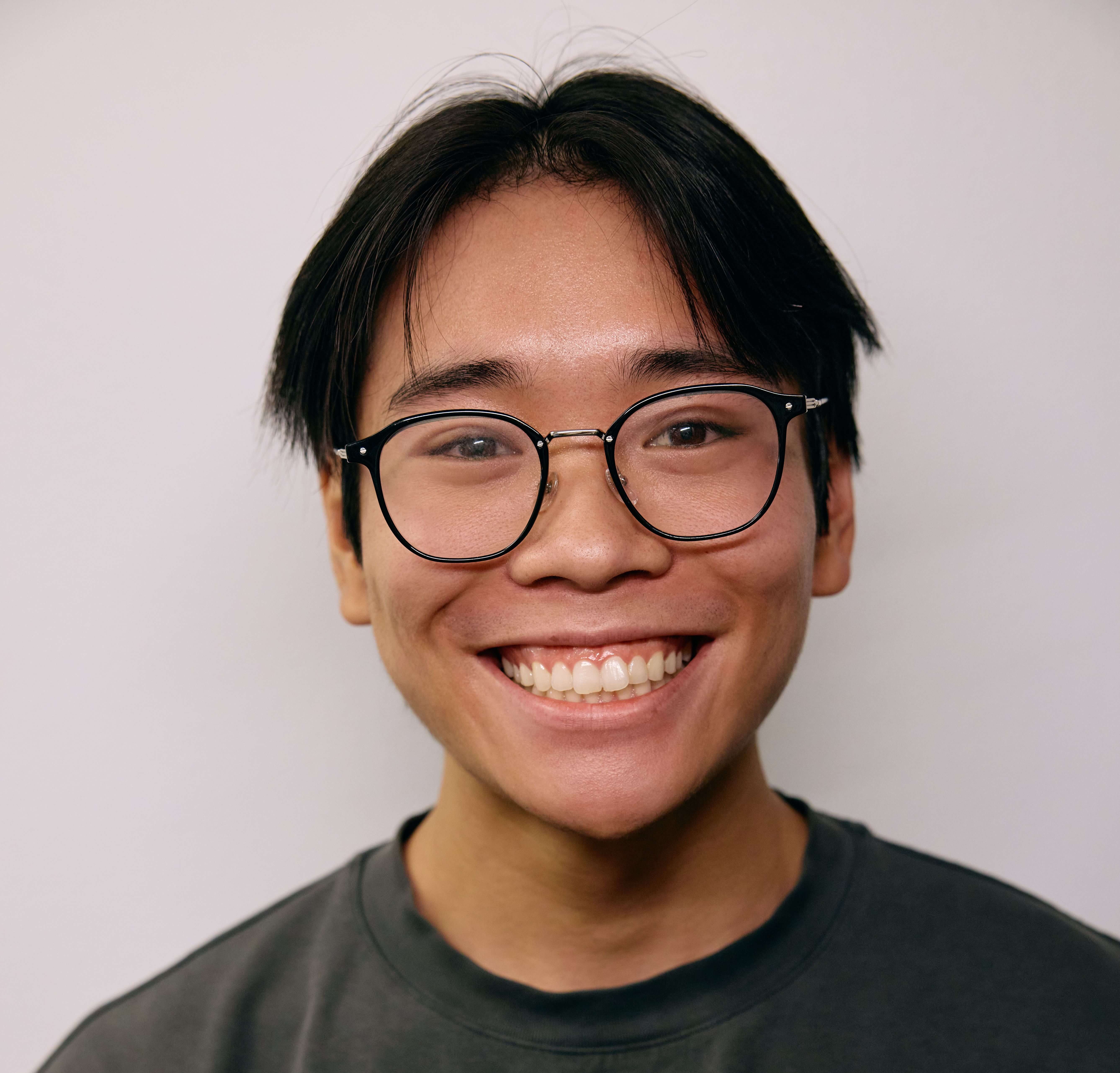 Smiling person with glasses, dark hair, wearing a dark shirt against a plain background. Their expression conveys happiness and friendliness.