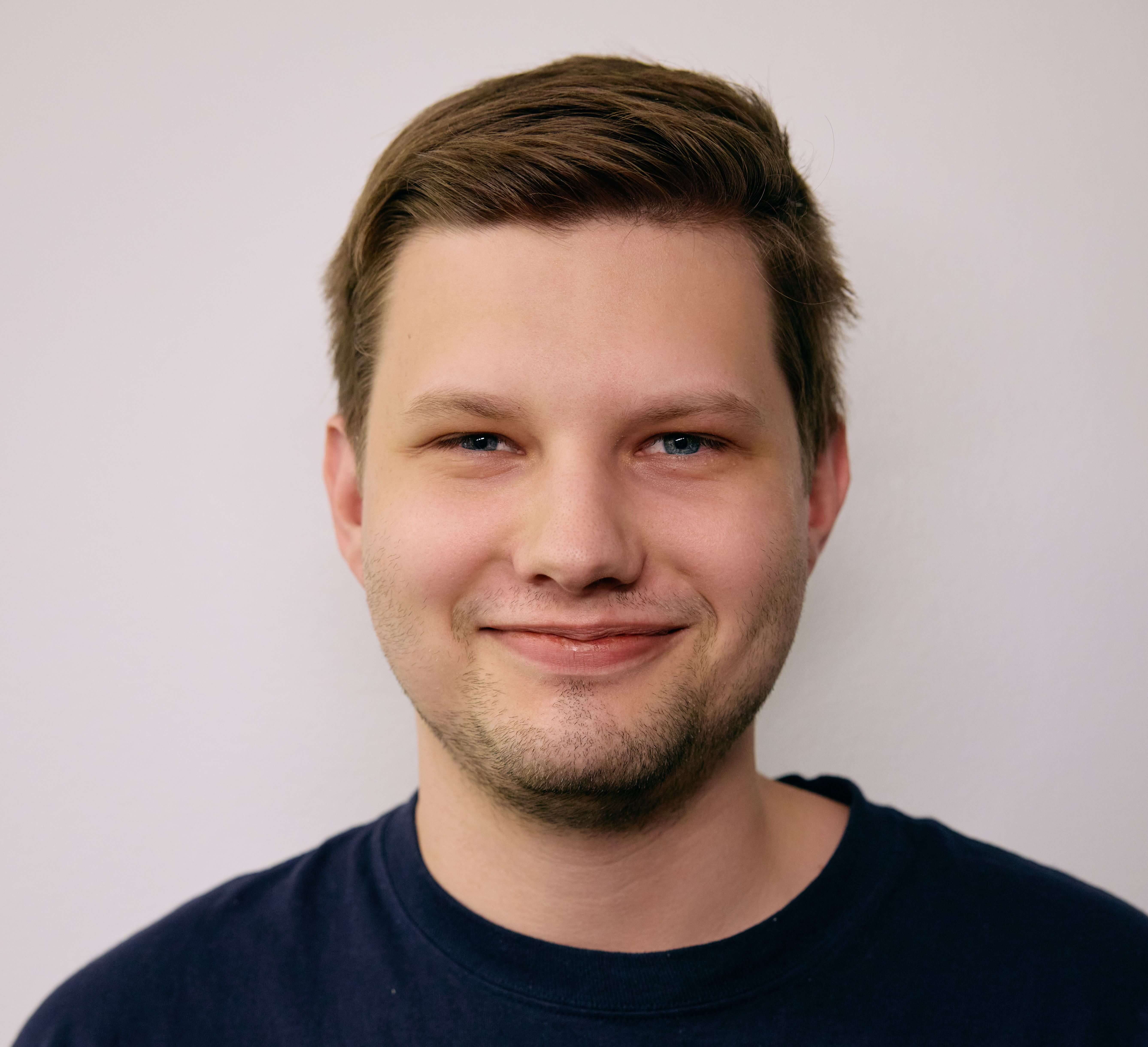 A smiling person with short brown hair and blue eyes is wearing a dark shirt against a plain light background, conveying a friendly and approachable demeanor.