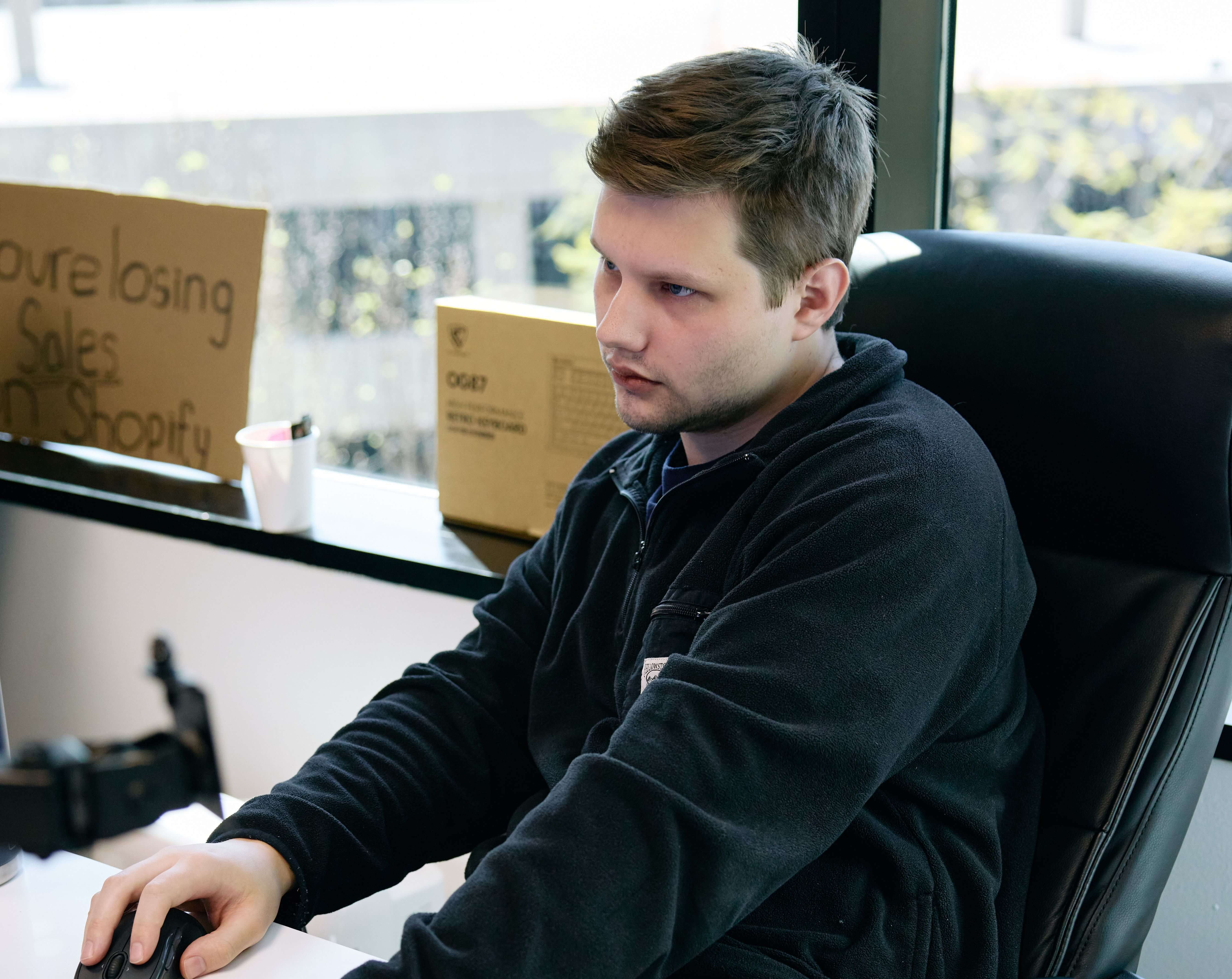 Man focused on a computer in a modern office, wearing a dark sweater. A cardboard sign by the window reads, "You're losing sales on Shopify."