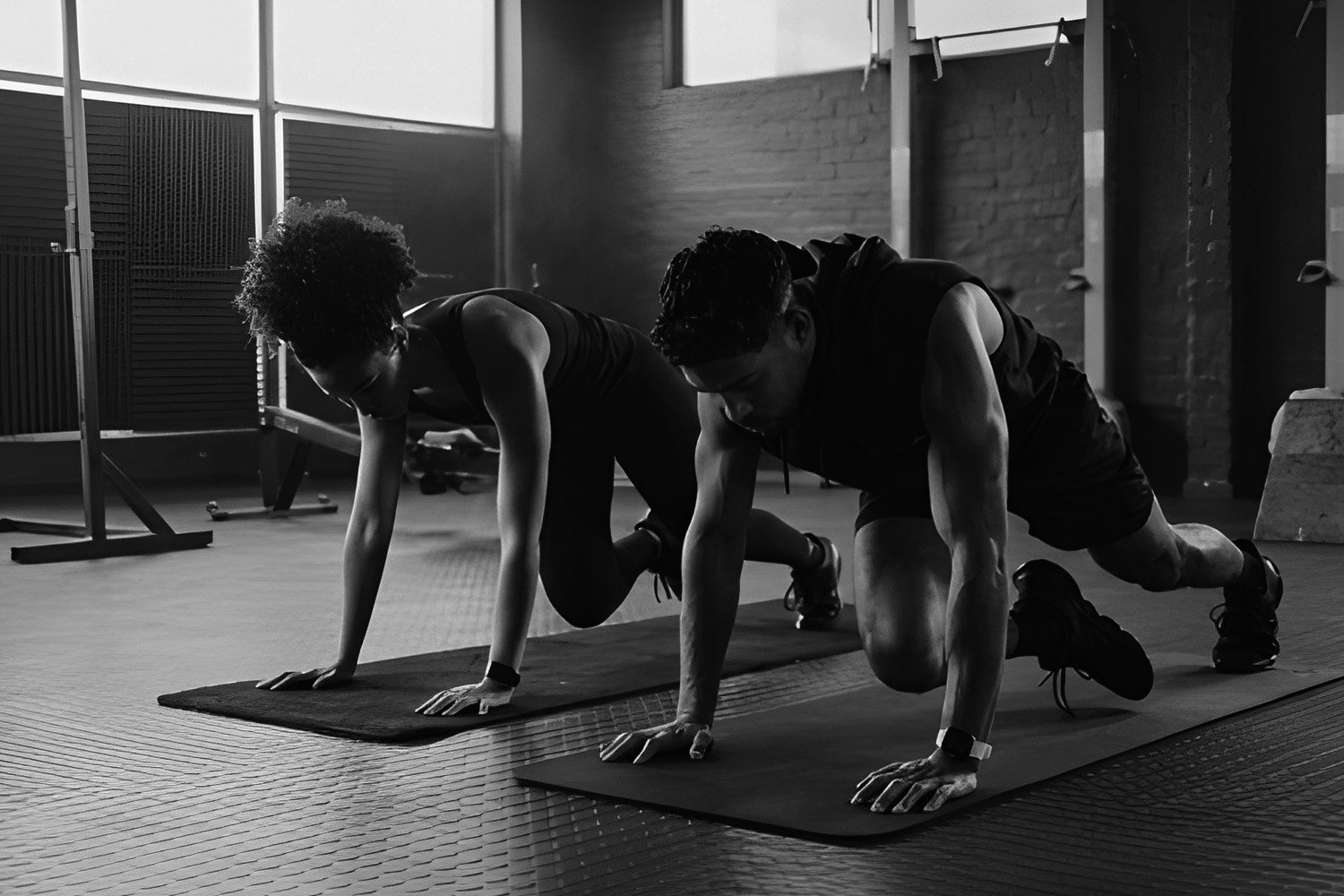 A man and woman in athletic wear performing mountain climber exercises on mats in a modern industrial-style gym.