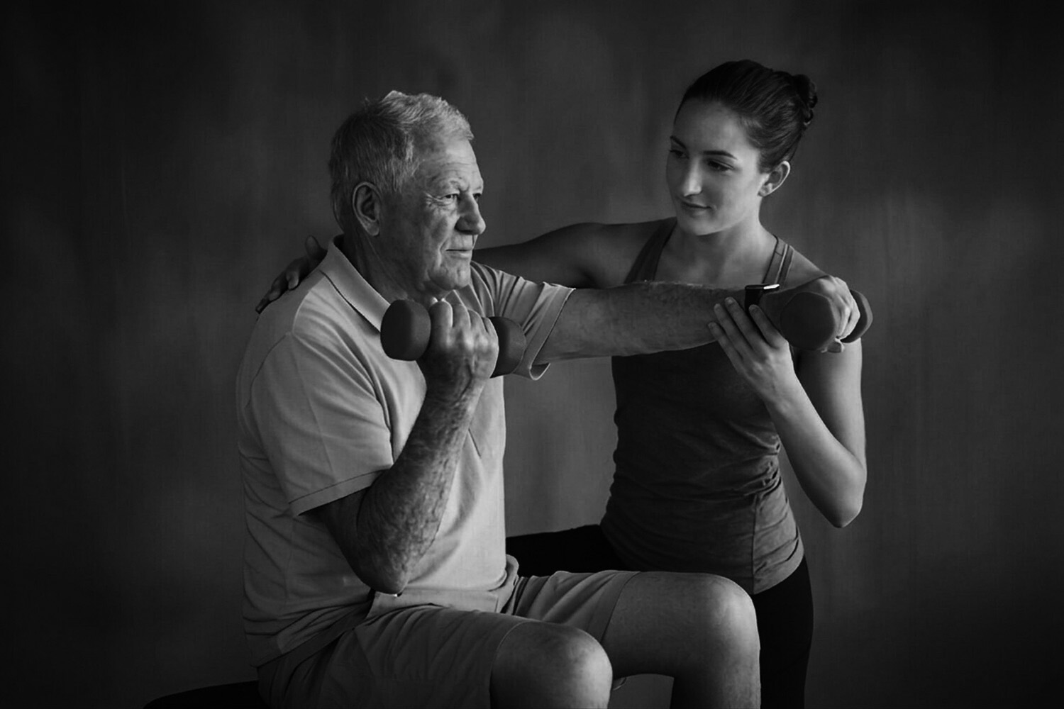 A senior man in a blue polo shirt performing a dumbbell exercise while a female trainer assists him.