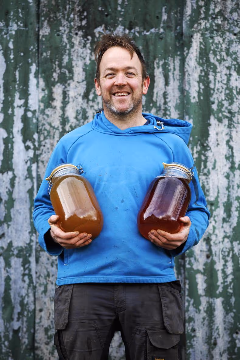Man in blue shirt holding two large jars of honey, standing in front of a weathered, green wall.