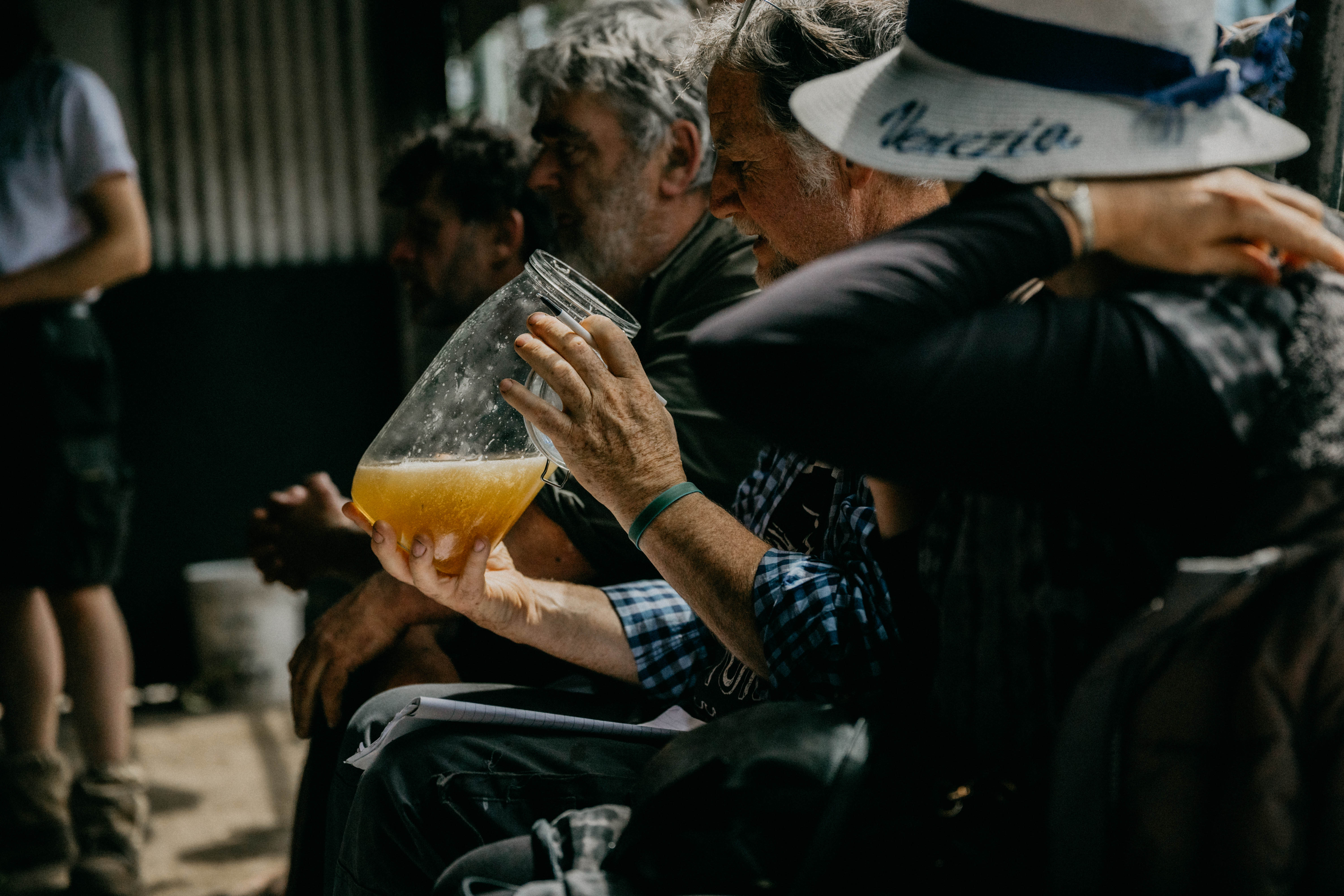 Man closely examines a jar with yellow liquid while sitting with others in a dimly lit room.
