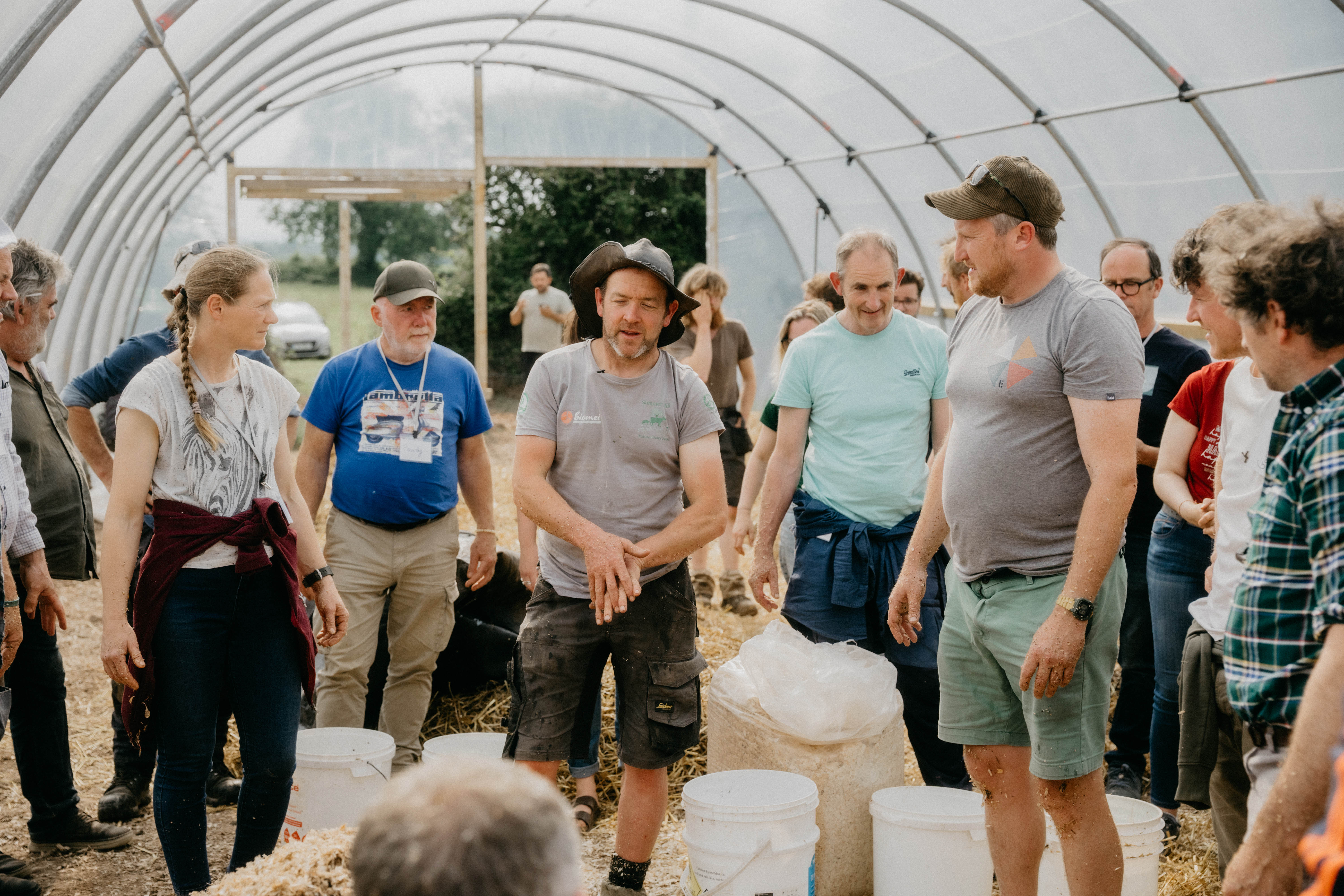 Group of people standing in a greenhouse listening to a man demonstrating with his hands.