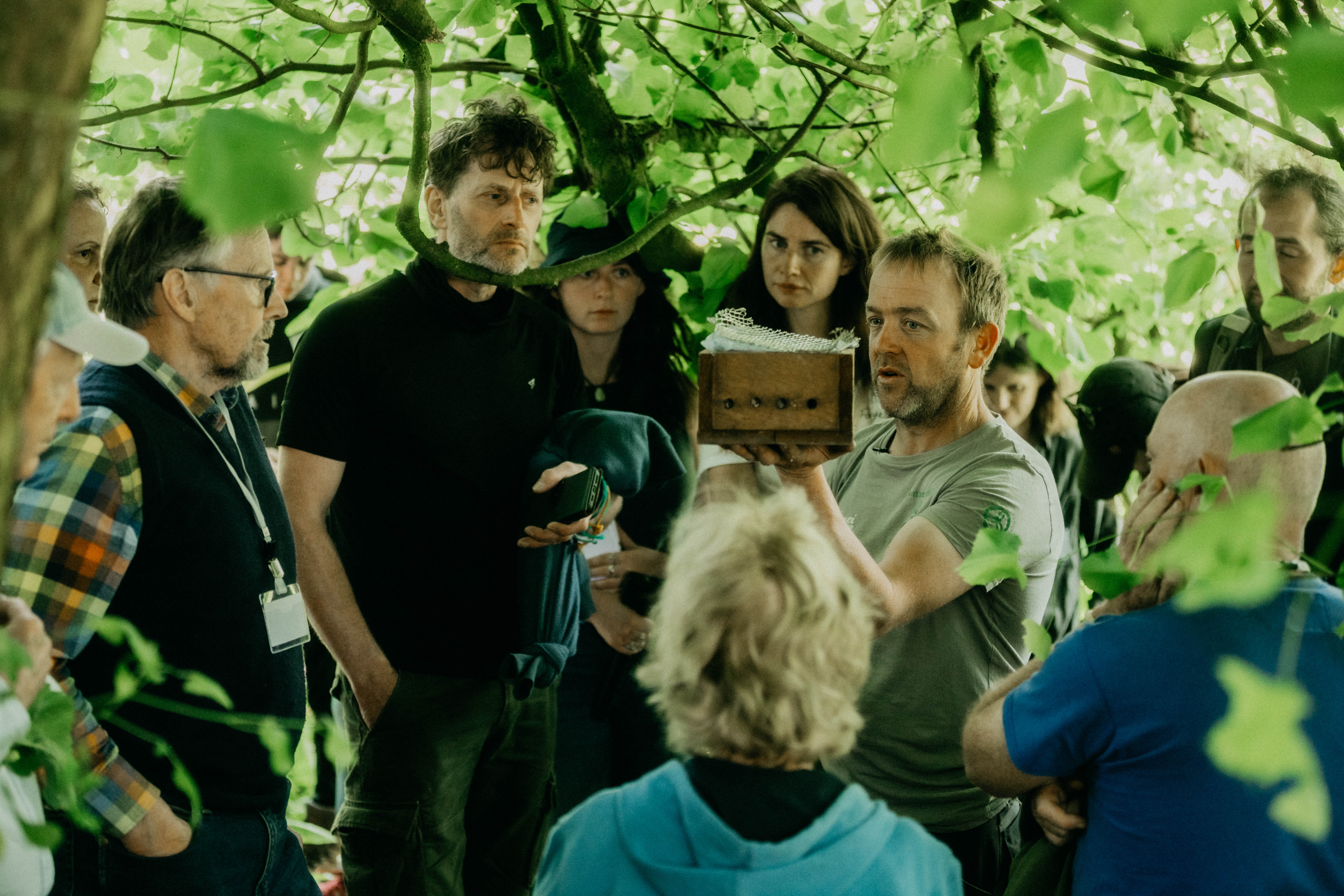 Man holding a wooden box, explaining to a group gathered closely under green leafy trees.