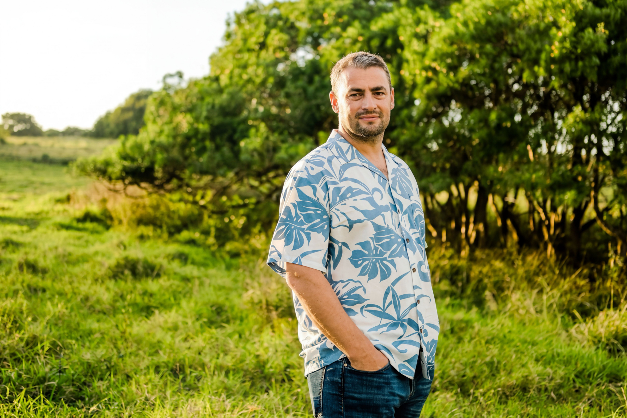 Man with short hair wearing a blue leaf-patterned shirt and jeans standing in grassy field near trees. Chris-Trump