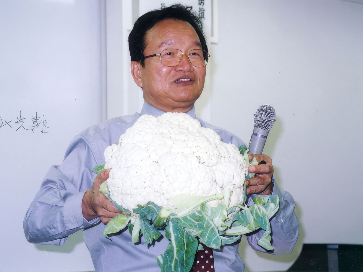 Man in glasses holding a large cauliflower and a microphone indoors.