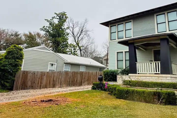 Side view of a two-story house with a porch, a fenced yard, and a nearby single-story garage. A freshly cut tree stump is visible in the lawn. Overcast sky in the background.