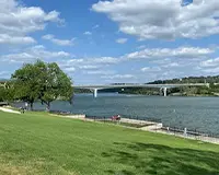 A serene riverside scene in Marble Falls Texas featuring a vivid green park lawn, a bridge spanning the water, and a partly cloudy blue sky.