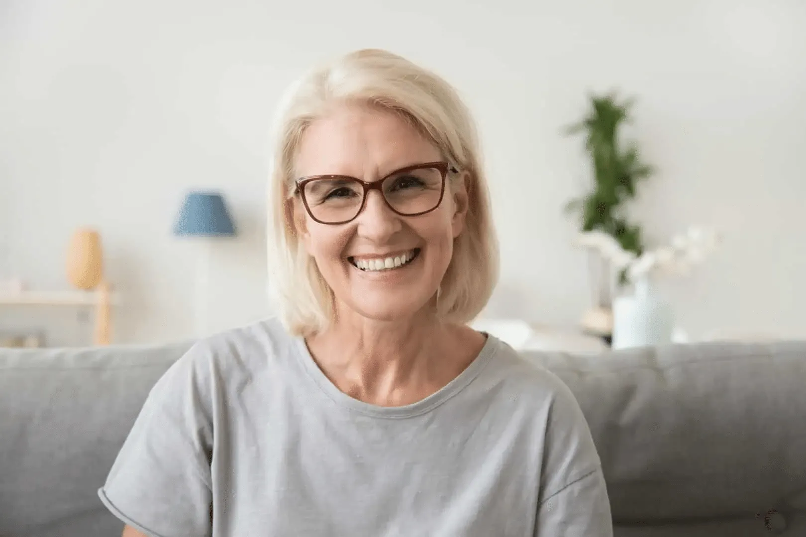 Smiling older woman with short blonde hair and glasses sitting on a grey couch in a bright room.