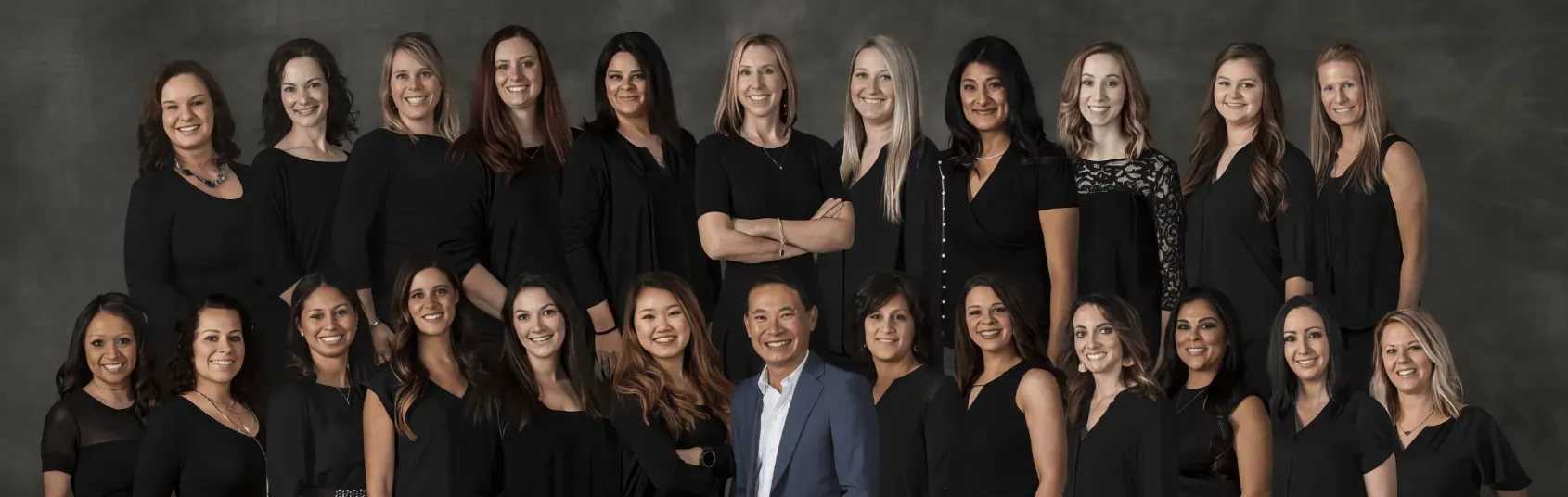 Group portrait of twenty women and one man dressed in black and gray standing and smiling against a dark gray background.