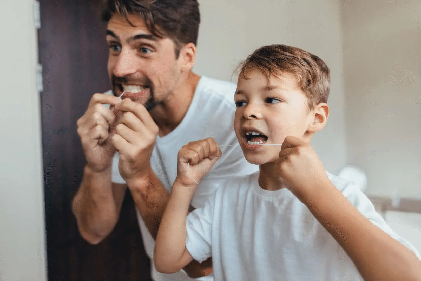 Young man and boy flossing their teeth together in a bathroom.